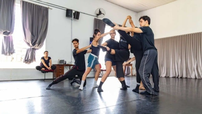 Grupo de jovens bailarinos ensaiando movimento contemporâneo em estúdio, com apoio mútuo na execução da pose e um instrutor sentado ao fundo observando.