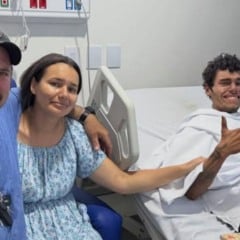 Jovem resgatado ap&oacute;s se perder na Serra do Japi aparece deitado em leito hospitalar ao lado de familiares, consciente e fazendo sinal de positivo.