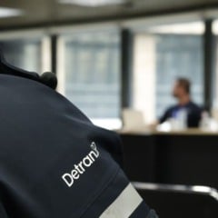 Servidor do Detran-SP de costas com uniforme oficial, enquanto pessoas participam de uma reunião ao fundo em ambiente administrativo.