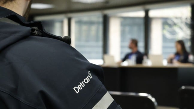 Servidor do Detran-SP de costas com uniforme oficial, enquanto pessoas participam de uma reunião ao fundo em ambiente administrativo.