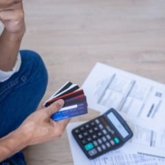 Homem sentado no ch&atilde;o segurando v&aacute;rios cart&otilde;es de cr&eacute;dito, com calculadora e contas espalhadas ao lado, representando preocupa&ccedil;&atilde;o com d&iacute;vidas e inadimpl&ecirc;ncia.