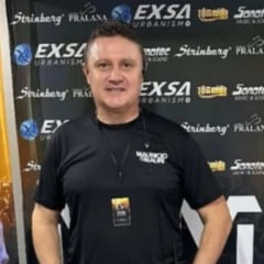 Homem sorridente, Douglas Riva, usando camiseta preta com o nome “Maurício & Mauri”, em frente a um painel com logomarcas de patrocinadores.