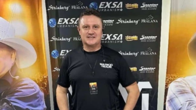 Homem sorridente, Douglas Riva, usando camiseta preta com o nome “Maurício & Mauri”, em frente a um painel com logomarcas de patrocinadores.