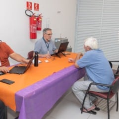 Homens atendem candidatos em uma mesa com computadores durante processo seletivo do programa Emprega+ no CRAS Sul, em Jundiaí.