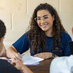 Recrutadora sorrindo enquanto entrevista dois jovens candidatos, segurando documentos sobre a mesa em um ambiente claro e simples.