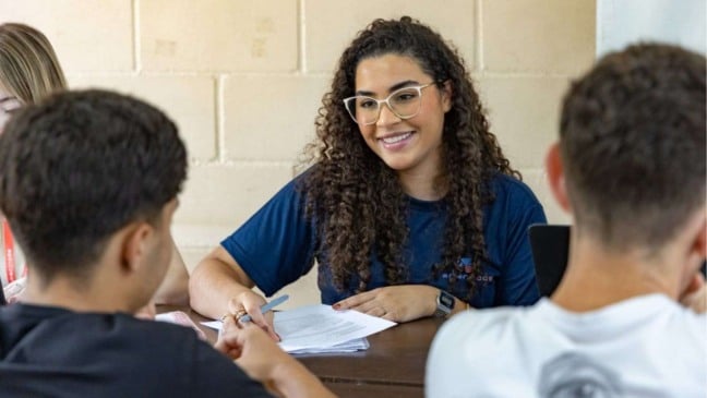 Recrutadora sorrindo enquanto entrevista dois jovens candidatos, segurando documentos sobre a mesa em um ambiente claro e simples.