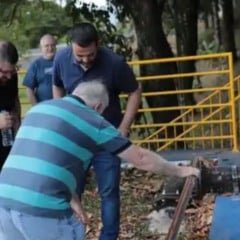 Homem manuseando a estrutura de uma adutora enquanto autoridades e participantes observam durante visita técnica em área externa, com grade de proteção amarela ao fundo.