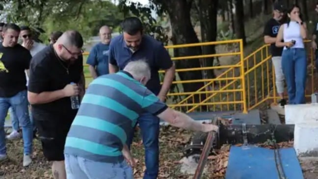 Homem manuseando a estrutura de uma adutora enquanto autoridades e participantes observam durante visita técnica em área externa, com grade de proteção amarela ao fundo.