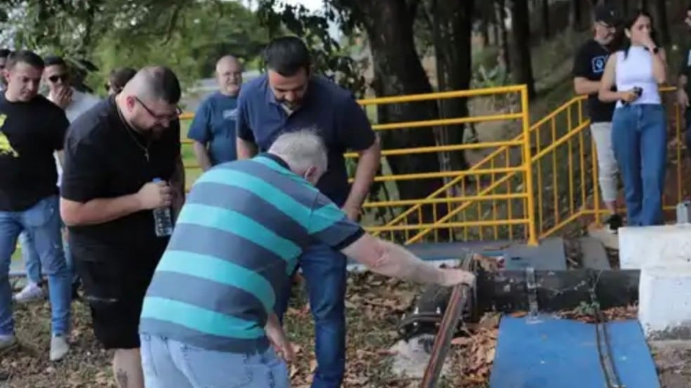 Homem manuseando a estrutura de uma adutora enquanto autoridades e participantes observam durante visita técnica em área externa, com grade de proteção amarela ao fundo. Homem manuseando a estrutura de uma adutora enquanto autoridades e participantes observam durante visita técnica em área externa, com grade de proteção amarela ao fundo.