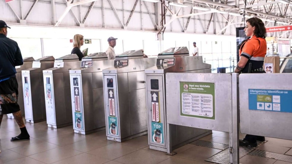 Catracas de acesso da estação da Linha 7-Rubi com passageiros circulando e agente de segurança uniformizada acompanhando o fluxo no interior da estação. Catracas de acesso da estação da Linha 7-Rubi com passageiros circulando e agente de segurança uniformizada acompanhando o fluxo no interior da estação.