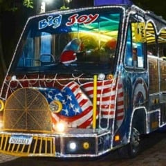 Ônibus do Expresso Natalino iluminado e decorado com luzes coloridas, exibindo detalhes temáticos e Papai Noel ao volante, estacionado à noite em uma rua arborizada.