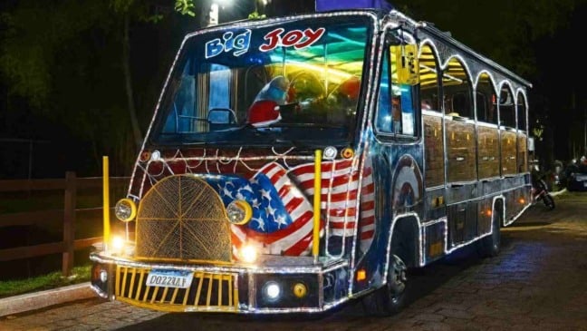 Ônibus do Expresso Natalino iluminado e decorado com luzes coloridas, exibindo detalhes temáticos e Papai Noel ao volante, estacionado à noite em uma rua arborizada.