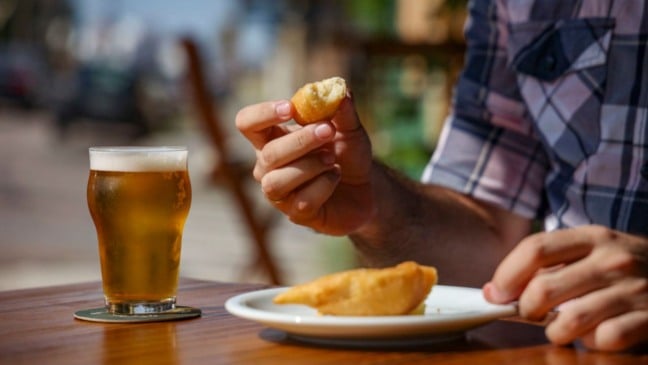 Homem segurando uma coxinha de queijo e, à mesa, um copo de cerveja artesanal gelada durante momento de degustação ao ar livre.