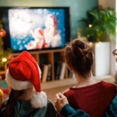 Fam&iacute;lia assiste a um filme de Natal na sala de estar decorada, com &aacute;rvore de Natal iluminada e crian&ccedil;a usando gorro natalino.
