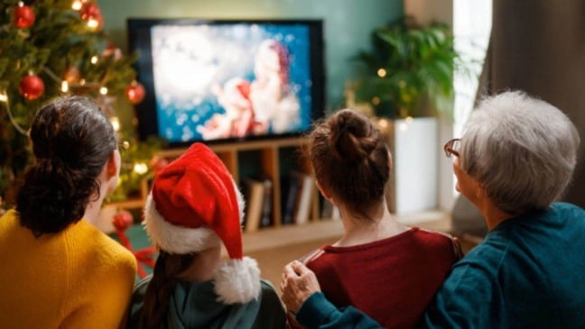 Família assiste a um filme de Natal na sala de estar decorada, com árvore de Natal iluminada e criança usando gorro natalino.
