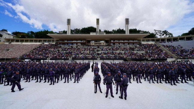 Panorama de um estádio com grande público nas arquibancadas e formação de centenas de policiais posicionados no centro durante cerimônia de formatura.