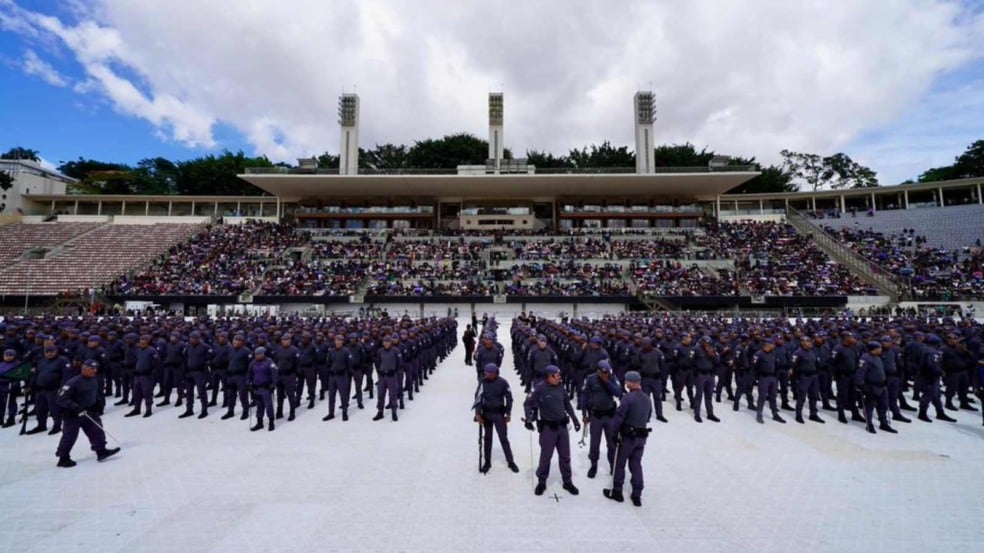 Panorama de um estádio com grande público nas arquibancadas e formação de centenas de policiais posicionados no centro durante cerimônia de formatura.