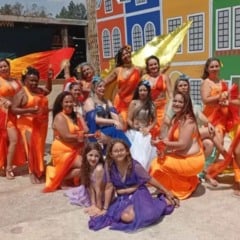Grupo de bailarinas de dan&ccedil;a do ventre posa sorrindo ao ar livre, com figurinos coloridos e leques, em cen&aacute;rio cultural decorado.