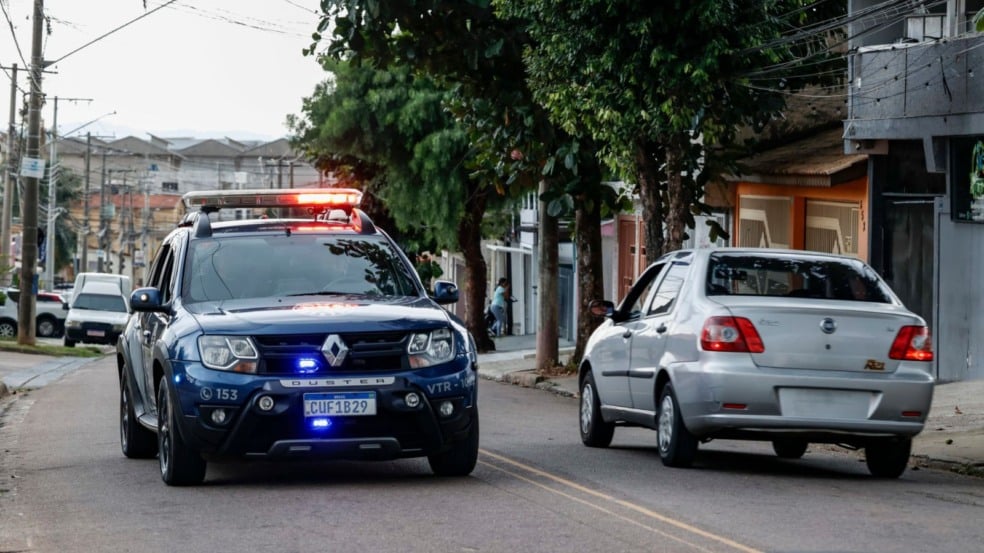 Viatura da Guarda Municipal de Jundiaí com giroflex ligado atrás de um carro prata em uma rua de bairro, com casas e pedestres ao fundo. Viatura da Guarda Municipal de Jundiaí com giroflex ligado atrás de um carro prata em uma rua de bairro, com casas e pedestres ao fundo.