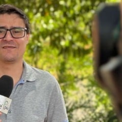 Homem de óculos segurando microfone da TVTEC Jundiaí enquanto grava uma reportagem ao ar livre, com vegetação desfocada ao fundo.