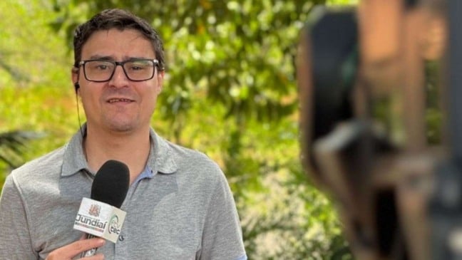 Homem de óculos segurando microfone da TVTEC Jundiaí enquanto grava uma reportagem ao ar livre, com vegetação desfocada ao fundo.