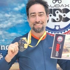 Barítono Guilherme Rosa segurando medalha e certificado após completar a prova BRM 1000 de Cabreúva, diante de painel do Audax ABC.