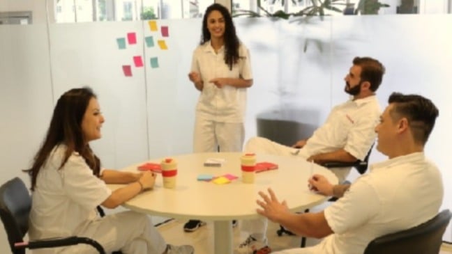 Grupo de profissionais em uniforme claro participa de reunião em sala moderna, com uma pessoa em pé apresentando ideias e post-its coloridos ao fundo.