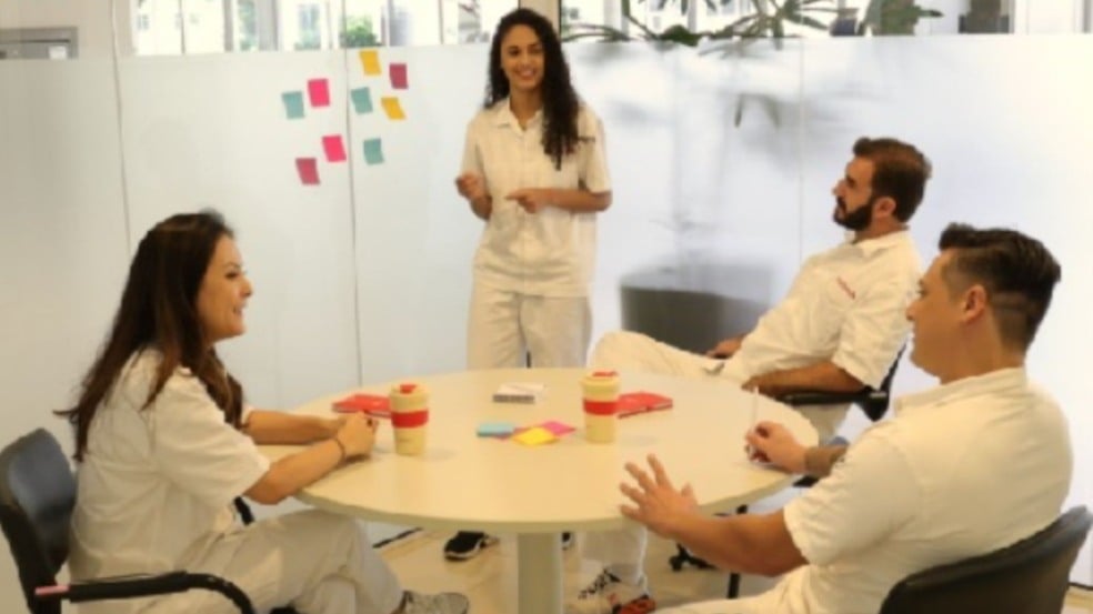 Grupo de profissionais em uniforme claro participa de reunião em sala moderna, com uma pessoa em pé apresentando ideias e post-its coloridos ao fundo. Grupo de profissionais em uniforme claro participa de reunião em sala moderna, com uma pessoa em pé apresentando ideias e post-its coloridos ao fundo.