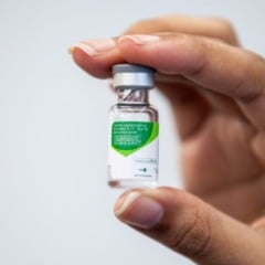 Mão segurando um frasco da vacina contra o HPV, com rótulo verde e branco em destaque.