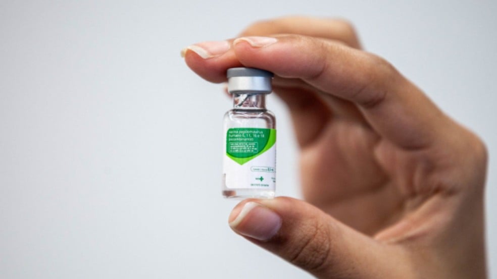 Mão segurando um frasco da vacina contra o HPV, com rótulo verde e branco em destaque. Mão segurando um frasco da vacina contra o HPV, com rótulo verde e branco em destaque.