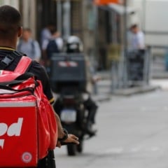 Entregador com mochila vermelha do iFood pedala pela rua enquanto outros motociclistas de delivery circulam ao redor em uma via urbana movimentada.