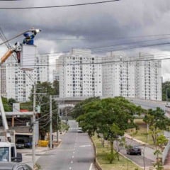 Trabalhador troca luminária de iluminação pública em poste da avenida, com prédios residenciais e via urbana ao fundo, em Jundiaí.