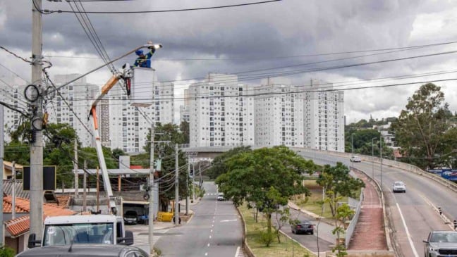 Trabalhador troca luminária de iluminação pública em poste da avenida, com prédios residenciais e via urbana ao fundo, em Jundiaí.