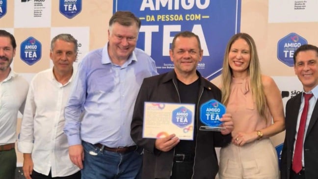 Grupo de seis pessoas posa sorrindo diante de um painel azul do Selo Amigo da Pessoa com TEA; ao centro, Edison Gonçalves segura o certificado e o troféu recebidos pela APAE de Jundiaí.