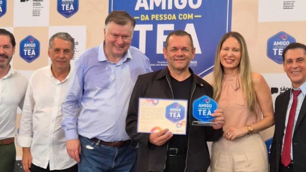 Grupo de seis pessoas posa sorrindo diante de um painel azul do Selo Amigo da Pessoa com TEA; ao centro, Edison Gonçalves segura o certificado e o troféu recebidos pela APAE de Jundiaí.