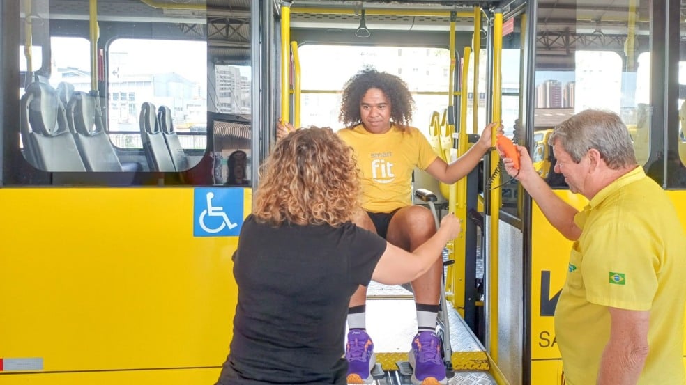 Pessoa participando de atividade de inclusão é erguida na plataforma elevatória de um ônibus acessível, acompanhada por dois orientadores no Terminal Vila Arens, em Jundiaí.