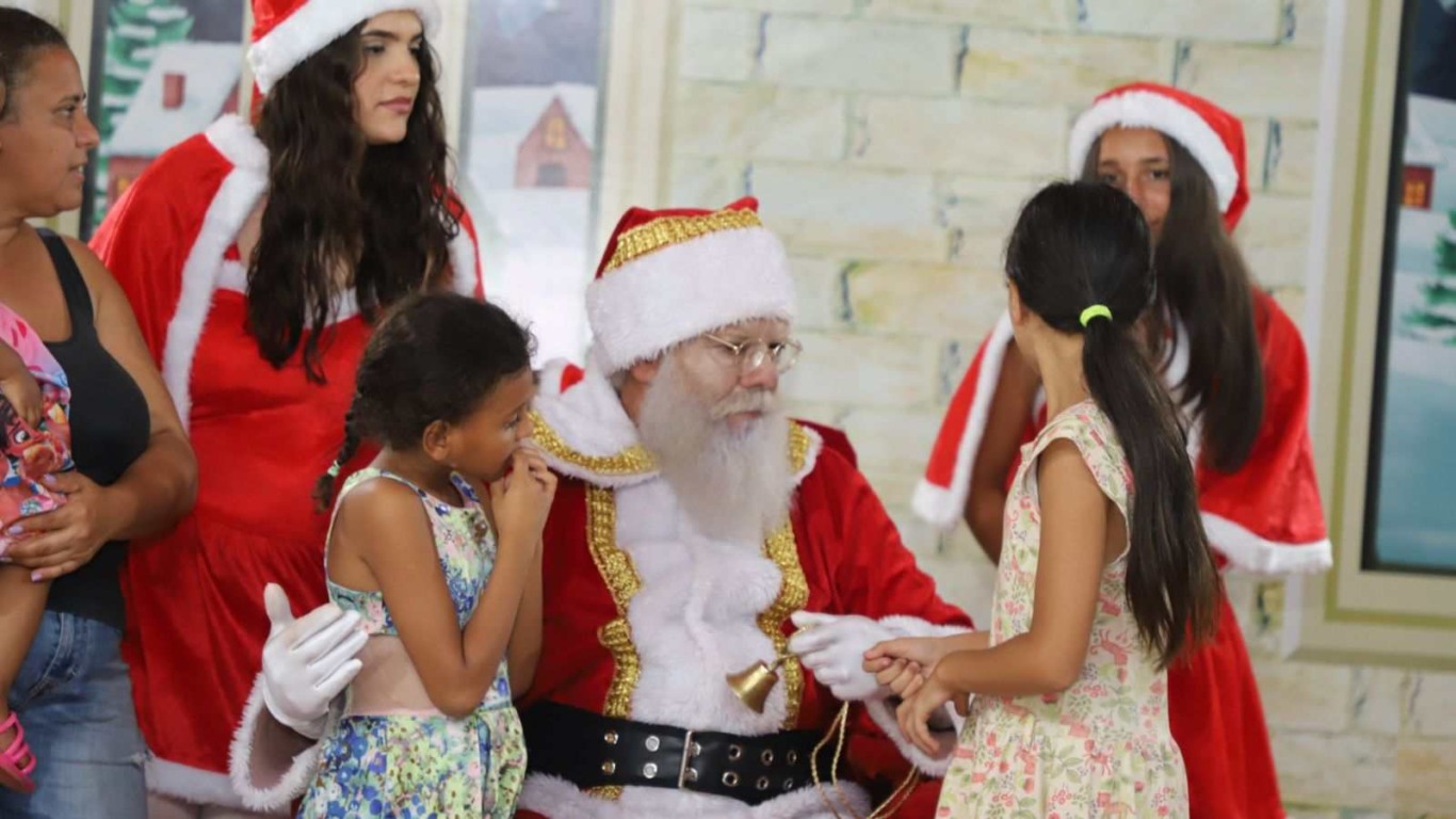 Crianças conversando com Papai Noel sentado, acompanhadas pela mãe e por ajudantes vestidas de Mamãe Noel, em ambiente decorado de Natal. Crianças conversando com Papai Noel sentado, acompanhadas pela mãe e por ajudantes vestidas de Mamãe Noel, em ambiente decorado de Natal.
