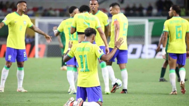 Jogador da Seleção Brasileira ajoelhado no gramado comemorando, com as mãos erguidas, enquanto outros atletas ao redor celebram usando o uniforme amarelo, azul e verde.
