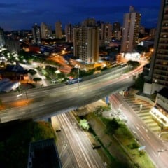 Vista a&eacute;rea noturna de Jundia&iacute; com avenida elevada iluminada, pr&eacute;dios residenciais e fluxo de carros nas ruas da cidade.