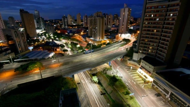 Vista aérea noturna de Jundiaí com avenida elevada iluminada, prédios residenciais e fluxo de carros nas ruas da cidade.
