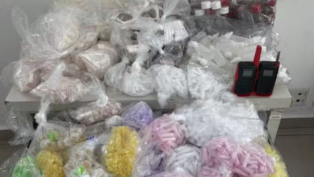Imagem com diversos sacos plásticos contendo drogas apreendidas — porções de maconha, cocaína, crack, haxixe, frascos de lança-perfume e dois rádios comunicadores organizados sobre uma mesa.