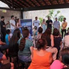 Grupo de moradores sentados ouvindo autoridades da Prefeitura e da DAE durante a entrega da nova rede de água no Loteamento São Jorge, em Jundiaí, em um espaço coberto, com banner da obra ao fundo.