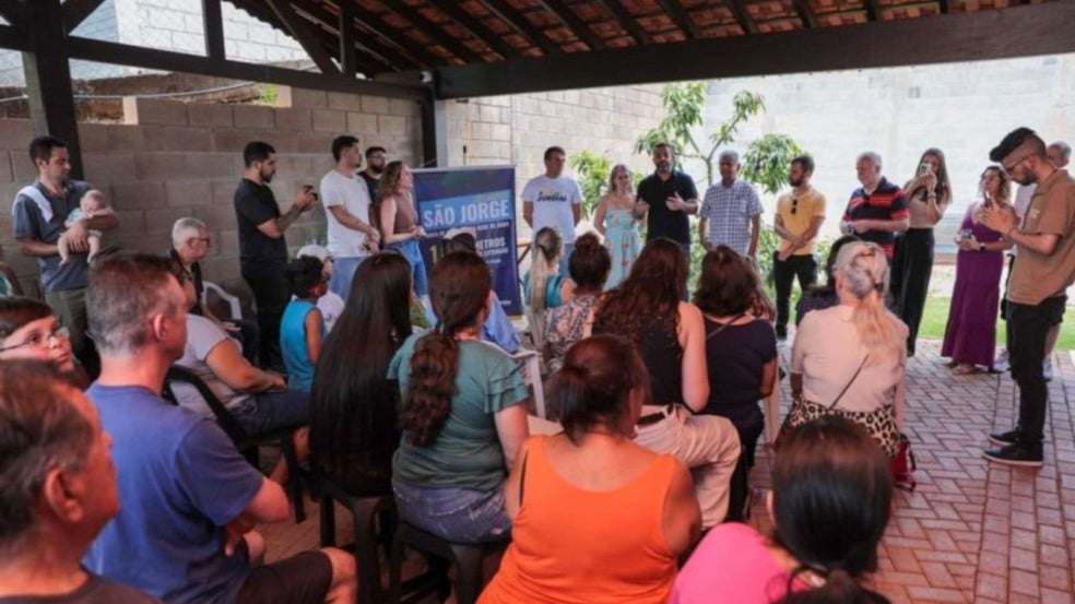 Grupo de moradores sentados ouvindo autoridades da Prefeitura e da DAE durante a entrega da nova rede de água no Loteamento São Jorge, em Jundiaí, em um espaço coberto, com banner da obra ao fundo.