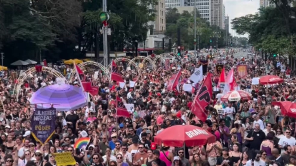 Multidão ocupa a Avenida Paulista durante a mobilização nacional “Mulheres Vivas”, com manifestantes segurando bandeiras, faixas e cartazes contra o feminicídio; prédios e decoração urbana aparecem ao fundo. Multidão ocupa a Avenida Paulista durante a mobilização nacional “Mulheres Vivas”, com manifestantes segurando bandeiras, faixas e cartazes contra o feminicídio; prédios e decoração urbana aparecem ao fundo.