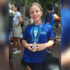 Catarina Di Fiore Cascioli, atleta mirim de Jundia&iacute;, sorri enquanto segura seu trof&eacute;u do Circuito Mirim 2025, usando a camiseta azul do Time Jundia&iacute; em um ambiente externo com outras pessoas ao fundo.