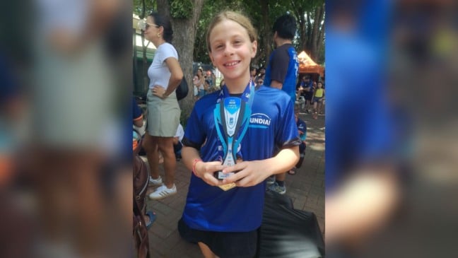 Catarina Di Fiore Cascioli, atleta mirim de Jundiaí, sorri enquanto segura seu troféu do Circuito Mirim 2025, usando a camiseta azul do Time Jundiaí em um ambiente externo com outras pessoas ao fundo.