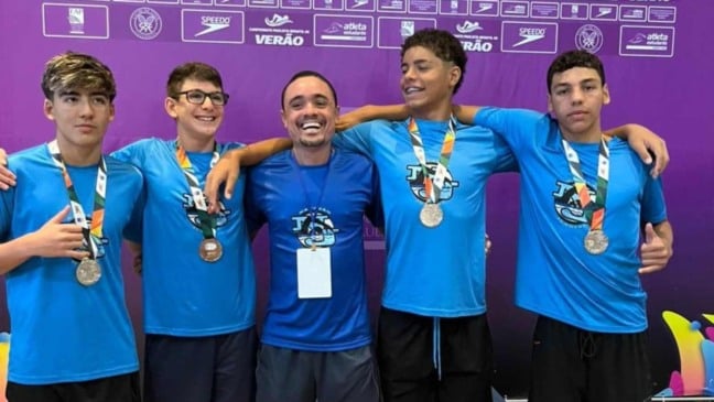 Atletas do Time Jundiaí e o técnico Marcelinho posando com medalhas em frente a painel roxo do campeonato.