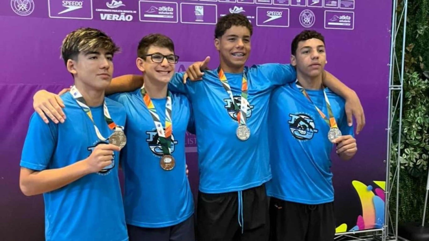 Quatro atletas do Time Jundiaí com camisetas azuis posam com medalhas diante de painel roxo do campeonato.