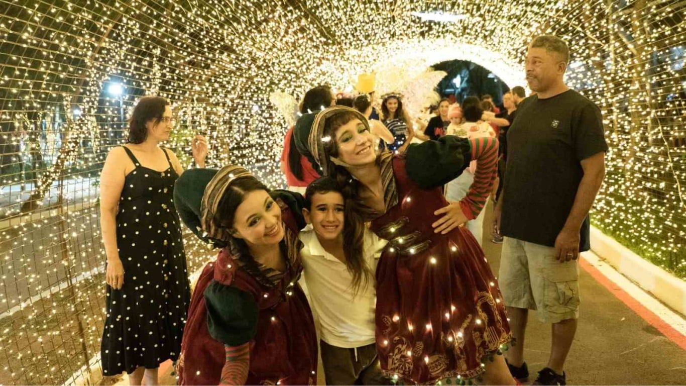 Duendes natalinos posam com uma criança dentro do Túnel Iluminado do Parque da Cidade, totalmente decorado com luzes douradas. Duendes natalinos posam com uma criança dentro do Túnel Iluminado do Parque da Cidade, totalmente decorado com luzes douradas.