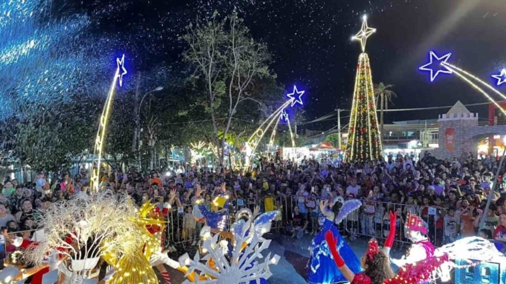 Multidão acompanha desfile natalino sob neve artificial, com artistas fantasiados e uma grande árvore de Natal iluminada ao fundo. Multidão acompanha desfile natalino sob neve artificial, com artistas fantasiados e uma grande árvore de Natal iluminada ao fundo.